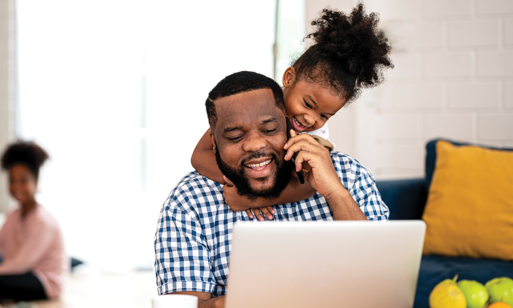 dad on the phone, looking at his laptop with his daughter playing on his shoulder